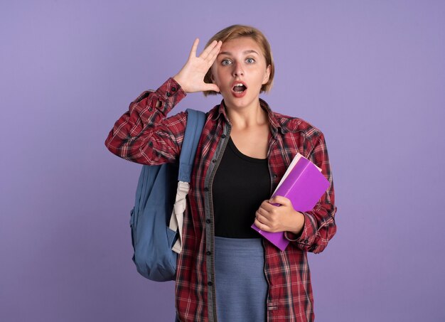 Überraschtes junges slawisches Studentenmädchen mit Rucksack legt Hand auf die Stirn, hält Buch und Notizbuch