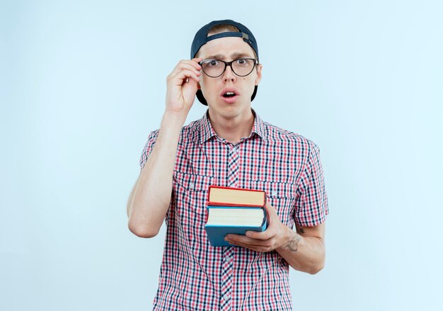 Überraschtes junges Studentenjungen, das Rückentasche und Brille und Kappe hält Bücher hält und Hand auf Brille lokalisiert auf weißer Wand setzt