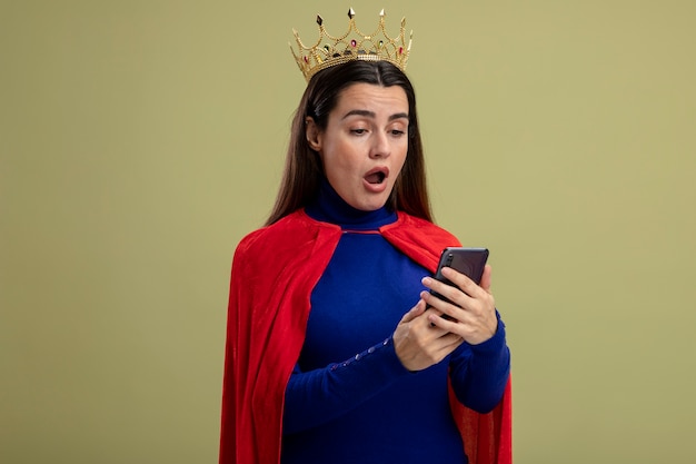 Überraschtes junges Superheldenmädchen, das Krone hält und Telefon lokalisiert auf olivgrün trägt