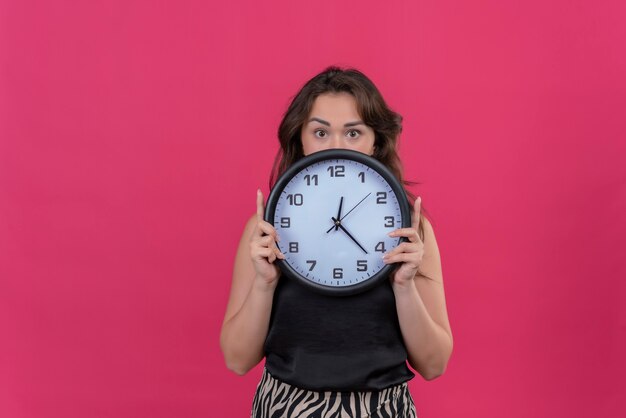 Überraschtes kaukasisches Mädchen, das schwarzes Unterhemd hält, das eine Wanduhr auf rosa Hintergrund hält