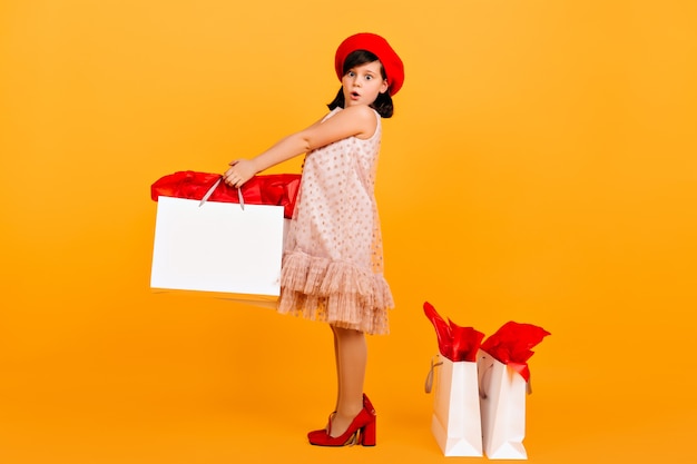 Überraschtes Kind, das in den Schuhen der Mutter aufwirft. erstauntes jugendliches Mädchen, das Einkaufstasche auf gelber Wand hält.