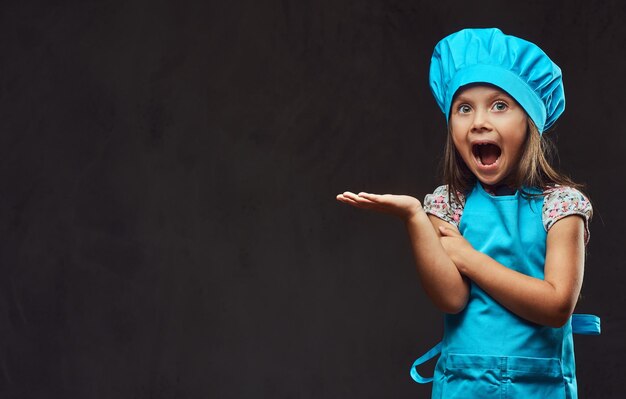 Überraschtes kleines Mädchen in blauem Koch posiert in einem Studio. Getrennt auf einem dunklen strukturierten Hintergrund.