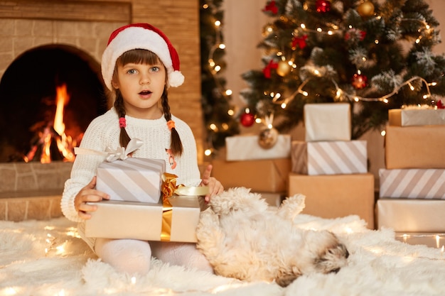 Überraschtes kleines Mädchen mit weißem Pullover und Weihnachtsmann-Hut, das mit Hund im festlichen Raum mit Kamin und Weihnachtsbaum posiert und Geschenkbox in Händen hält.