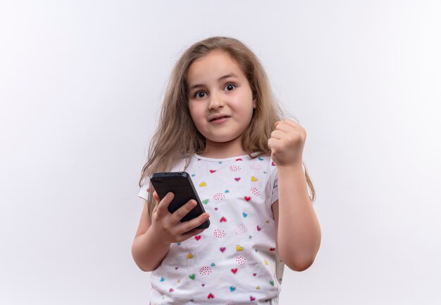 Überraschtes kleines Schulmädchen, das weißes T-Shirt hält Telefon hält und Faust auf lokalisiertem weißem Hintergrund erhebt