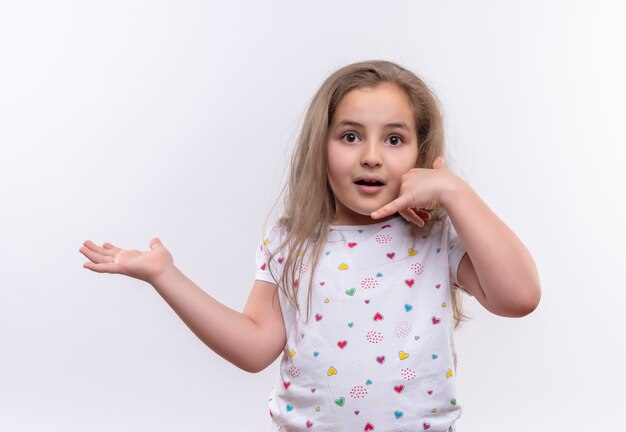 Überraschtes kleines Schulmädchen, das weißes T-Shirt trägt, das Anrufgeste auf lokalisiertem weißem Hintergrund zeigt