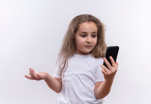 Überraschtes kleines Schulmädchen, das weißes T-Shirt trägt, das auf Telefon auf lokalisiertem weißem Hintergrund schaut