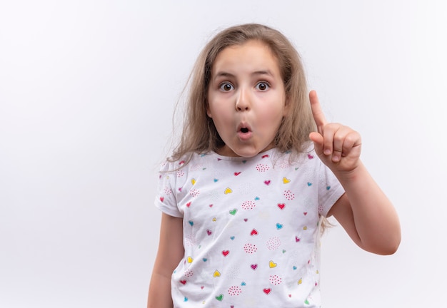 Überraschtes kleines Schulmädchen, das weißes T-Shirt trägt, zeigt auf lokalisierten weißen Hintergrund