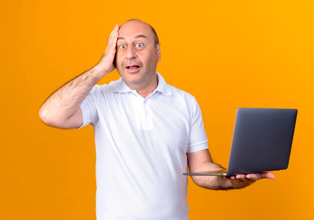 Überraschtes lässiges reifes Mann, das Laptop hält und Hand auf Stirn lokalisiert auf gelber Wand setzt