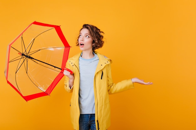 Überraschtes Mädchen im Mantel, der oben schaut und Regenschirm hält. schockierte junge Dame mit Sonnenschirm isoliert auf leuchtend orange Wand.