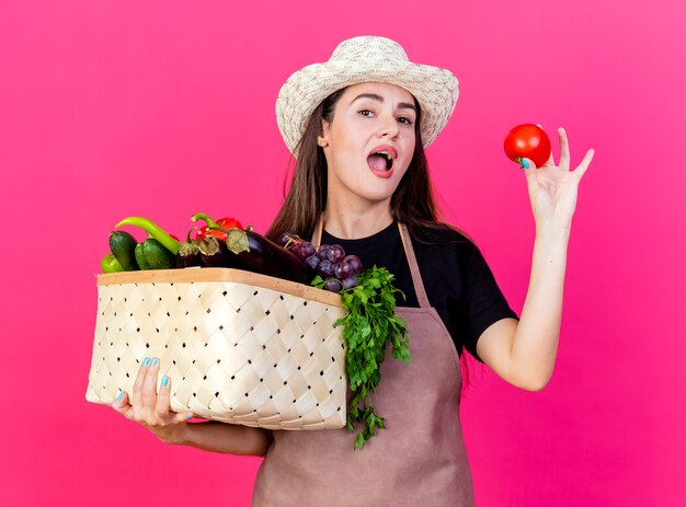 Überraschtes schönes Gärtnermädchen in der Uniform, die Gartenhut hält Gemüsekorb mit Tomate lokalisiert auf rosa Hintergrund