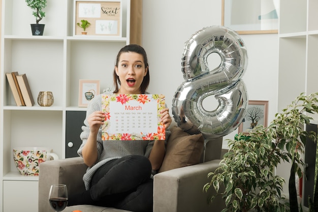 Überraschtes schönes Mädchen am glücklichen Frauentag, der den Kalender hält, der auf einem Sessel im Wohnzimmer sitzt