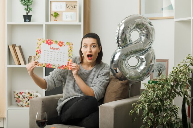 Überraschtes schönes Mädchen am glücklichen Frauentag, der den Kalender hält, der auf einem Sessel im Wohnzimmer sitzt