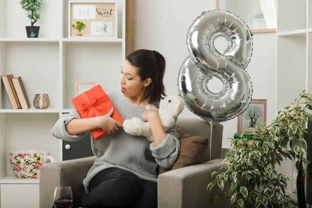 Überraschtes schönes Mädchen am glücklichen Frauentag, der gegenwärtig mit Teddybär auf einem Sessel im Wohnzimmer sitzt und schaut