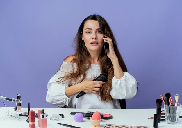 Überraschtes schönes Mädchen sitzt am Tisch mit Make-up-Werkzeugen hält Haarkamm, der am Telefon spricht, lokalisiert auf lila Wand