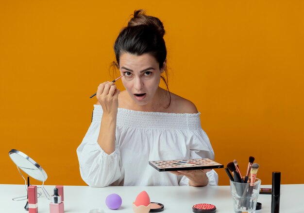 Überraschtes schönes Mädchen sitzt am Tisch mit Make-up-Werkzeugen hält Lidschatten-Palette Anwendung Lidschatten mit Make-up-Pinsel isoliert auf orange Wand