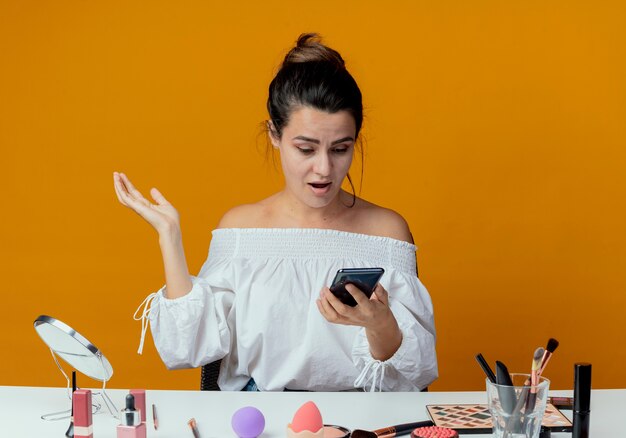 Überraschtes schönes Mädchen sitzt am Tisch mit Make-up-Werkzeugen hält und schaut auf Telefon isoliert auf orange Wand