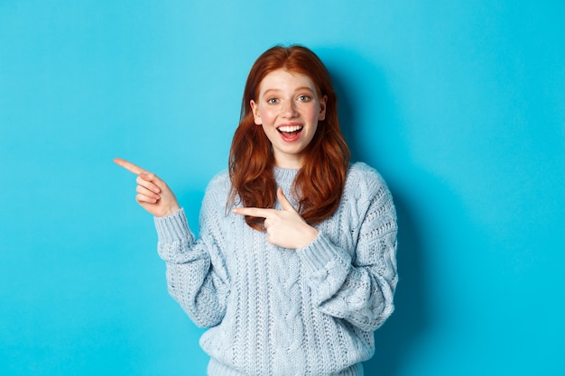 Überraschtes Teenager-Mädchen mit roten Haaren und Sommersprossen, das mit den Fingern nach links auf das Logo zeigt und lächelt, Werbung zeigt und auf blauem Hintergrund steht.