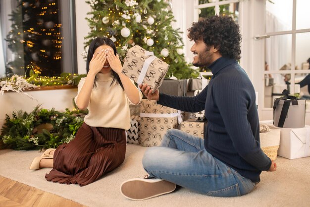 Überraschung. Junge erwachsene Frau in festlicher Kleidung, die Augen schließt und einen lächelnden indischen Mann hält, der das Geschenk seitlich zur Kamera hält, die auf dem Boden in der Nähe des Weihnachtsbaums in einem beleuchteten Raum sitzt