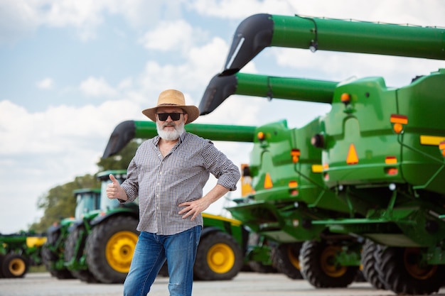 Berufsbauer mit einem modernen traktor, mähdrescher auf einem feld im sonnenlicht bei der arbeit. landwirtschaft, ausstellung, maschinen, pflanzenbau. älterer mann in der nähe seiner maschine.