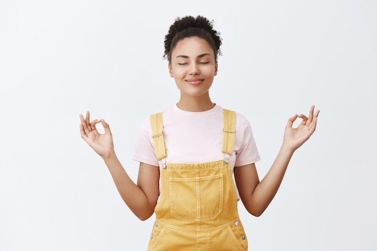 Kostenlose Foto beruhige dich und lass den stress im inneren los. ruhige und niedliche gut aussehende dunkelhäutige frau in gelben trendigen overalls, die mit zen-geste hände auf beiden seiten hält, meditiert oder yoga macht