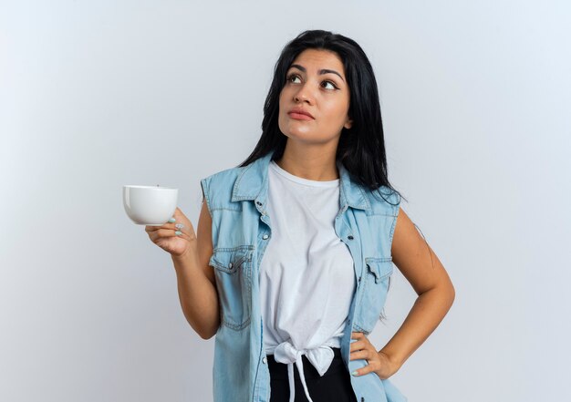 Überzeugte junge kaukasische Frau hält Tasse, die auf Seite schaut