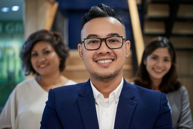 Überzeugter asiatischer Geschäftsmann, der für Kamera und weibliche Kollegen hinten stehen lächelt