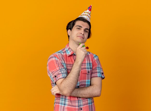 Überzeugter gutaussehender kaukasischer Mann mit Geburtstagsmütze hält Partypfeife