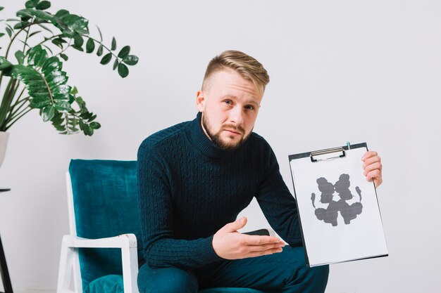 Überzeugter junger männlicher Psychologe, der auf dem Lehnsessel zeigt rorschach-inkblot Testpapier sitzt