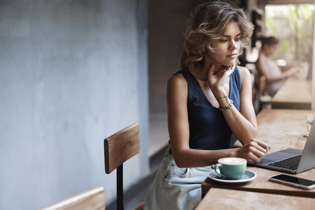 Beschäftigte attraktive junge moderne Geschäftsfrau bereiten Projektleseartikel vor sitzen in der Nähe von Fenster Cafétisch trinken Kaffee Denken Blick Laptop Display Studentin tutor Vorkehrungen treffen zukünftige Lektion