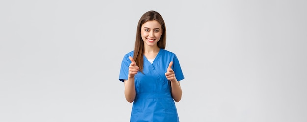 Beschäftigte im Gesundheitswesen verhindern Virusversicherung und Medizinkonzept Begeisterte lächelnde Ärztin in blauen Scrubs, die sich mit Pistolen zeigt und Kunden in die Klinik einlädt