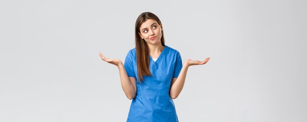 Beschäftigte im Gesundheitswesen verhindern Virusversicherung und Medizinkonzept Unentschlossene und ahnungslose attraktive Ärztin, Krankenschwester in blauen Scrubs, die mit den Händen seitlich zuckt und grinsend wegschaut