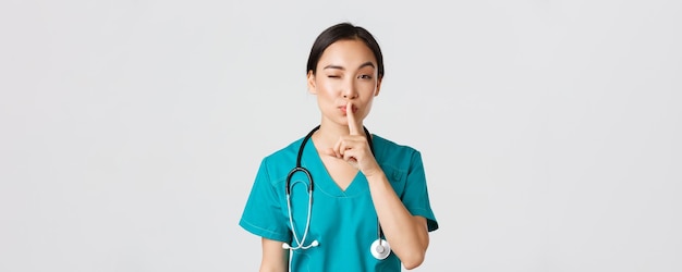 Beschäftigte im Gesundheitswesen, Virusprävention, Konzept der Quarantänekampagne. Freche süße asiatische Ärztin, Ärztin, die kokett schweigt und zwinkert, hat ein Geheimnis, bereitet eine Überraschung vor und bittet, ruhig zu bleiben