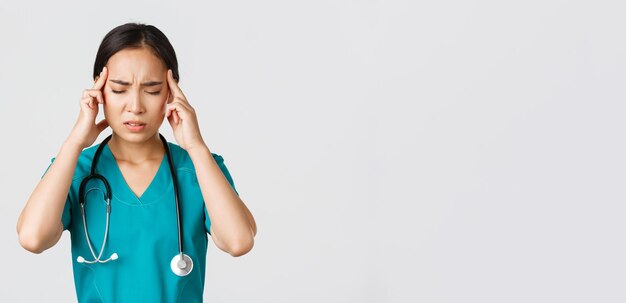 Beschäftigte im Gesundheitswesen, Virusprävention, Konzept der Quarantänekampagne. Nahaufnahme eines überarbeiteten und müden asiatischen Arztes, einer Krankenschwester, die Kopfschmerzen hat, beunruhigt aussieht, weißer Hintergrund steht.