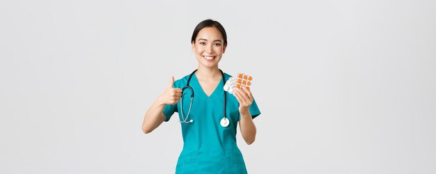 Beschäftigte im Gesundheitswesen, Virusprävention, Konzept der Quarantänekampagne. Zuversichtlich lächelnde asiatische Krankenschwester, Arzt in Scrubs empfehlen Medikamente, zeigen Daumen hoch und Pillen, weißer Hintergrund