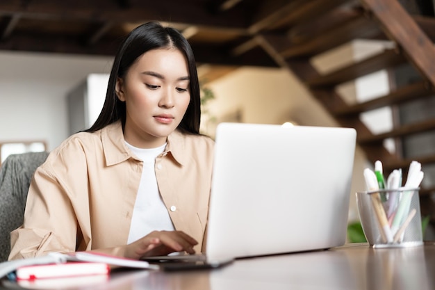 Beschäftigte junge asiatische frau arbeitet von zu hause mit laptop am tisch sitzend mit büchern und bleistiften tippt...