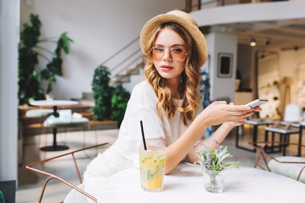 Beschäftigte junge Frau in der trendigen Brille und im Weinlesehut, die auf Anruf warten, während Sie im gemütlichen Café sitzen