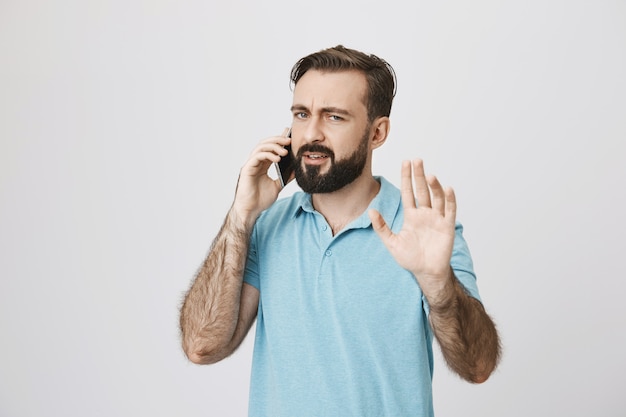 Beschäftigter bärtiger Kerl, der am Telefon spricht, Hand hebt und wartet