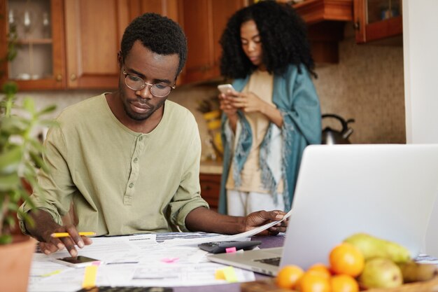 Beschäftigter ernsthafter afrikanischer Mann, der Handy benutzt, während Familienausgaben berechnet und Papierkram erledigt