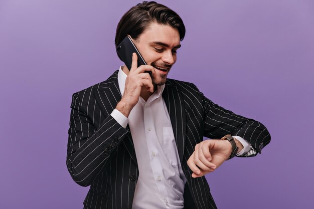 Beschäftigter junger Herr mit brünetten Haaren, weißem Hemd und schwarzem gestreiftem Anzug, der am Telefon spricht und auf die Uhr schaut und vor violettem Hintergrund lächelt Geschäftskonzept