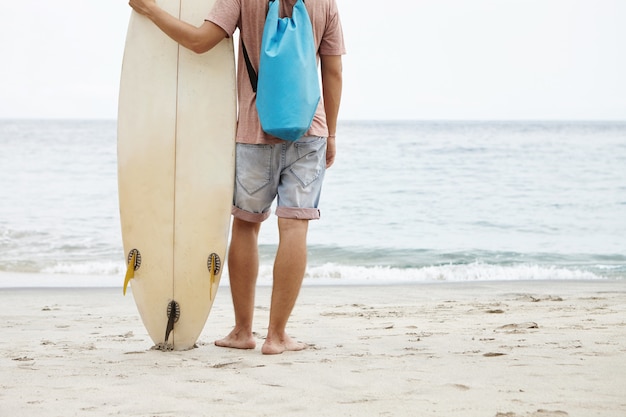 Beschnittene Ansicht des jungen Mannes mit blauem Rucksack, der auf Sandstrand steht und blaues Meerwasser betrachtet