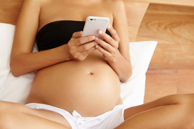 Beschnittener Schuss der jungen Frau, die Baby beim Surfen im Internet auf dem Handy erwartet
