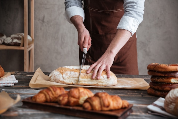 Beschnittenes Bild des Bäckers des jungen Mannes schnitt das Brot.