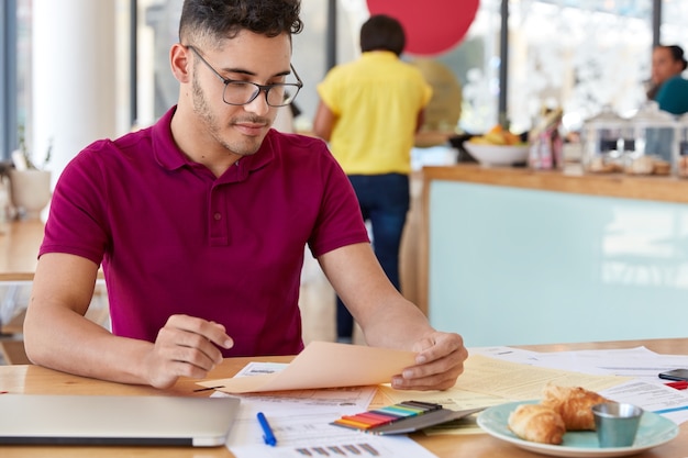 Beschnittenes Bild des Finanzberichts der männlichen Freiberuflerstudien, macht Projekt, gekleidet in Freizeitkleidung, sitzt am Schreibtisch mit bunten Aufklebern, Croissants, trägt lässiges Outfit. Papierkram-Konzept