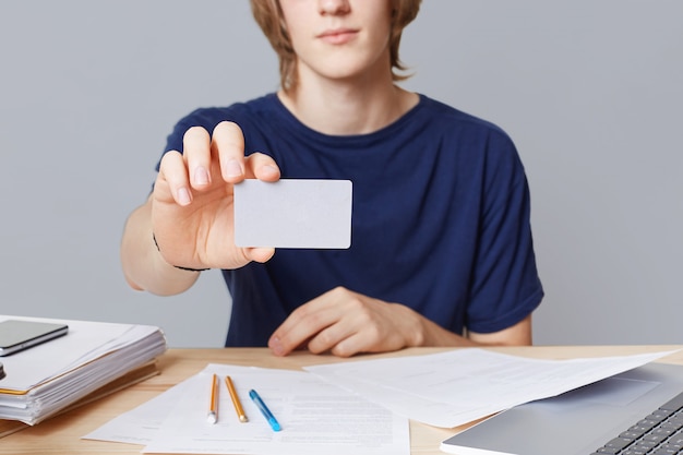 Beschnittenes Bild des lässig gekleideten jungen männlichen Unternehmers hält Karte mit blnk Kopienraum, sitzt auf Arbeitstisch, mit Papieren umgeben, über grauer Wand isoliert. Geschäftsmann hält Visitenkarte