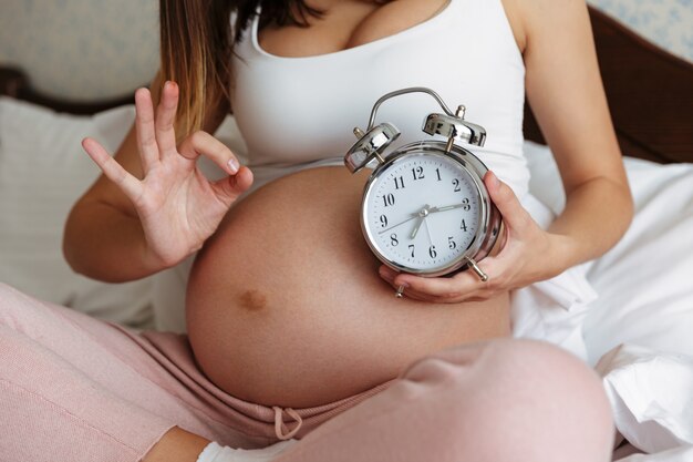 Beschnittenes Foto der schwangeren Frau, die Wecker hält
