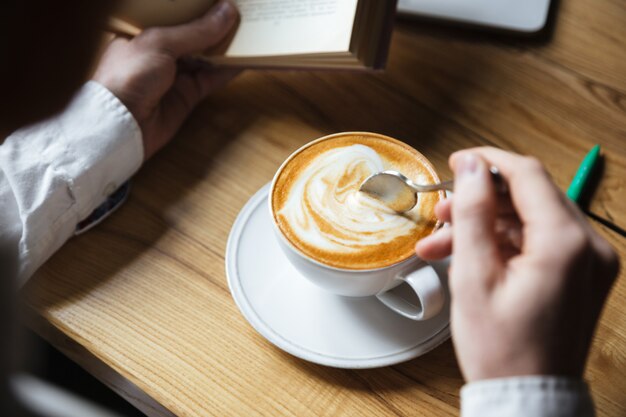 Beschnittenes Foto des Mannes im weißen Hemd, das Kaffee beim Lesen des Buches rührt