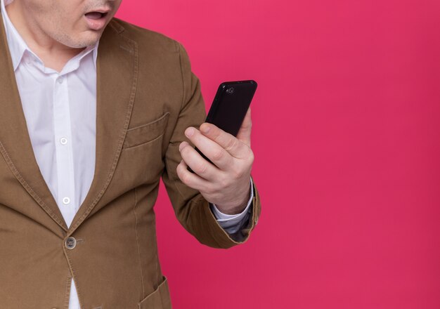 Beschnittenes Foto des Mannes mittleren Alters im Anzug, der Handy hält, das es erstaunt ansieht