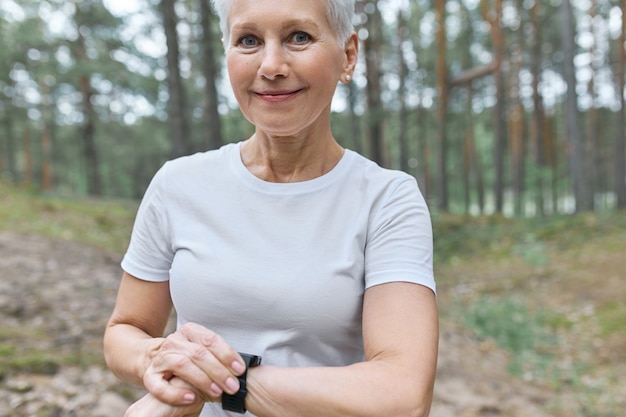 Beschnittenes Porträt der schönen Frau mittleren Alters im weißen T-Shirt, das intelligente Uhr an ihrem Handgelenk anpasst