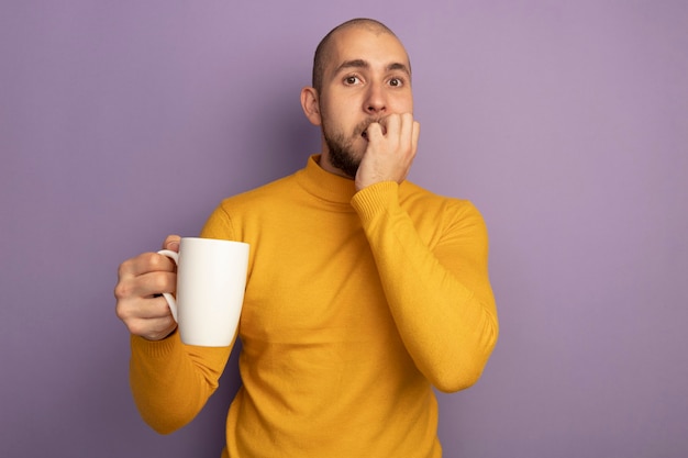 Besorgt, geradeaus schauend junger gutaussehender junger Mann, der Tasse Tee beißt, beißt Nägel lokalisiert auf Purpur mit Kopienraum