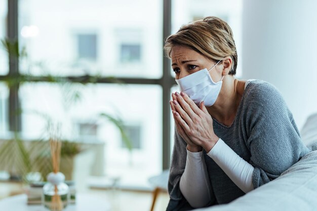 Besorgte Frau, die mit gefalteten Händen betet, während sie eine schützende Gesichtsmaske trägt und sich während einer Coronavirus-Pandemie traurig fühlt